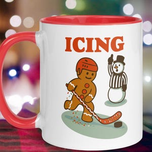 May include: White ceramic mug with a red interior and handle. The mug features a gingerbread man playing hockey with a snowman referee. The word "ICING" is printed in red above the illustrations. A fun, festive design for the holiday season.