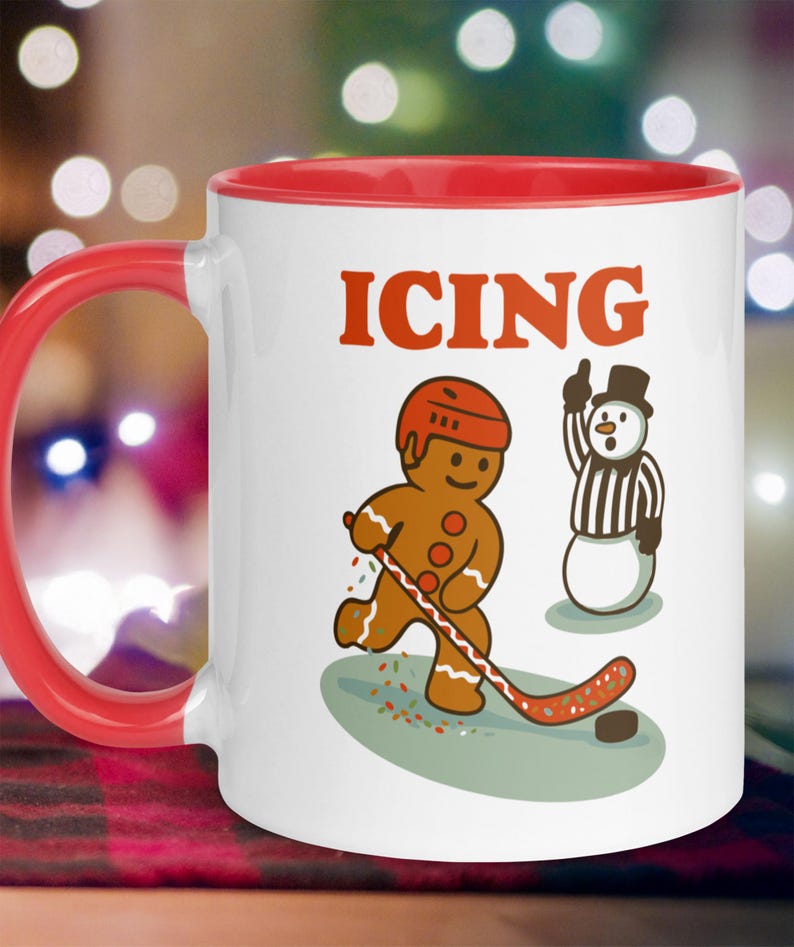 May include: White ceramic mug with a red interior and handle. The mug features a gingerbread man playing hockey with a snowman referee. The word "ICING" is printed in red above the illustrations. A fun, festive design for the holiday season.