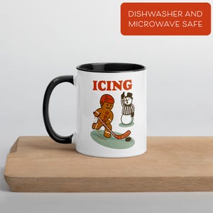 May include: White ceramic mug with a black interior and handle. The mug features a cartoon gingerbread man playing hockey with the word "ICING" in red. A snowman referee is also depicted. The mug is labeled "Dishwasher and Microwave Safe".