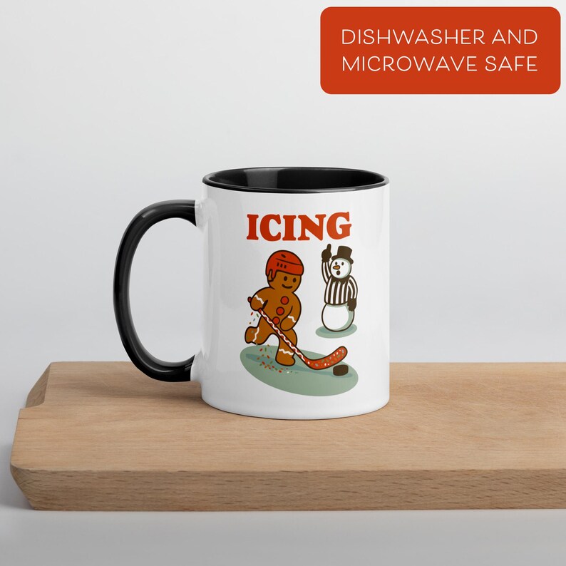 May include: White ceramic mug with a black interior and handle. The mug features a cartoon gingerbread man playing hockey with the word "ICING" in red. A snowman referee is also depicted. The mug is labeled "Dishwasher and Microwave Safe".