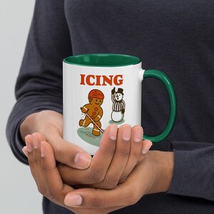 May include: White ceramic mug with a green interior and handle, featuring a gingerbread man playing hockey and a snowman referee. The word "ICING" is printed in red above the illustrations. The mug is held in both hands.