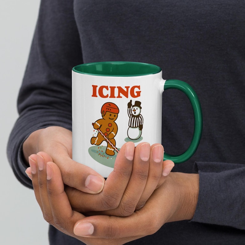 May include: White ceramic mug with a green interior and handle, featuring a gingerbread man playing hockey and a snowman referee. The word "ICING" is printed in red above the illustrations. The mug is held in both hands.