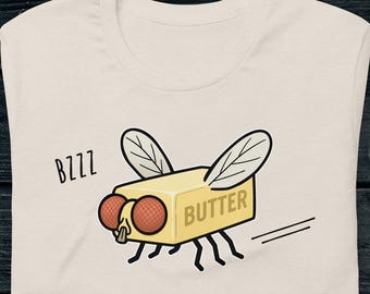 Può includere: T-shirt color crema con un'illustrazione umoristica di una mosca di burro. Il disegno mostra un panetto di burro con caratteristiche da mosca, tra cui ali, occhi e zampe. La parola "BUTTER" è stampata sul burro e "BZZZ" è stampato a sinistra.