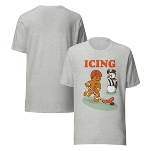 May include: Light gray t-shirt with a front graphic of a gingerbread man playing hockey, the word "ICING" in orange, and a snowman referee. The back of the shirt is plain.