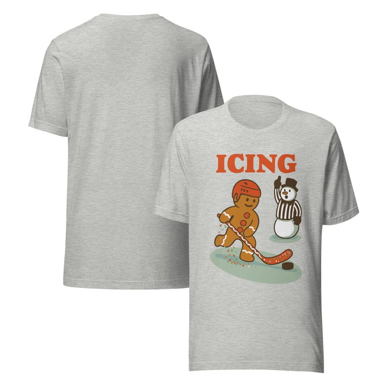 May include: Light gray t-shirt with a front graphic of a gingerbread man playing hockey, the word "ICING" in orange, and a snowman referee. The back of the shirt is plain.