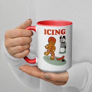 May include: White ceramic mug with a red interior and handle. The mug features a gingerbread man playing hockey with a snowman referee and the word "ICING" in red letters.