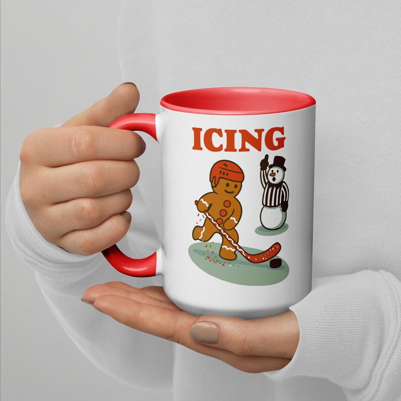 May include: White ceramic mug with a red interior and handle. The mug features a gingerbread man playing hockey with a snowman referee and the word "ICING" in red letters.