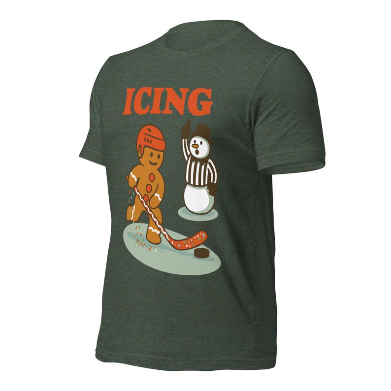 May include: Dark green t-shirt featuring a gingerbread man playing hockey against a snowman referee. The word "ICING" is printed in orange above the illustration. The gingerbread man wears a helmet and holds a hockey stick.