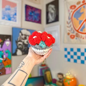 May include: A handmade crochet flower with red, white, gray, and teal accents. The flower is held in a hand and appears to be a decorative item. The crochet flower has a textured appearance and is likely made of yarn.