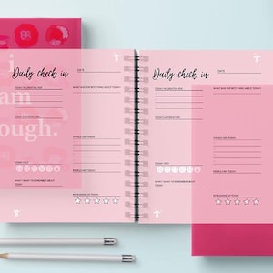 May include: A pink and red spiral bound journal with the text "Daily check in" and "I am enough." on the cover. The inside pages have a pink background with lines for writing and a section for rating your day.