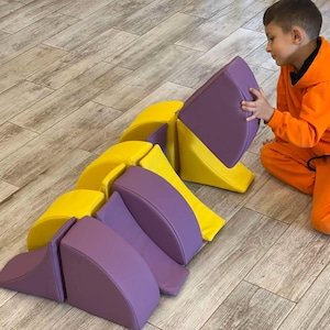 May include: A set of colorful foam blocks in yellow and purple. The blocks are arranged on a wooden floor. The blocks are in various shapes, including curved and angled pieces, suitable for building and play.