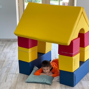 Puede incluir: Una colorida casa de juegos construida con grandes bloques de espuma blanda. La casa tiene un techo amarillo, paredes rojas y amarillas y bloques de base azules. Un niño está dentro de la casa de juegos, descansando sobre una almohada.