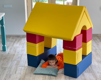 May include: A colorful playhouse constructed from large, soft foam blocks. The house features a yellow roof, red and yellow walls, and blue base blocks. A child is inside the playhouse, resting on a pillow.