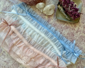 May include: Three ruffled, sheer fabric pouches in pastel pink, white, and blue, each tied with a matching ribbon bow. A large pink crystal and two smaller white crystals are visible, along with a bundle of dried flowers.
