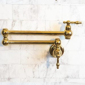 Op de afbeelding: Een goudkleurige, aan de muur gemonteerde potvulkraan met een dubbel scharnierende arm om het bereik te vergroten. De kraan heeft een decoratieve hendel en tuit en is gemonteerd op een wit baksteenpatroon.
