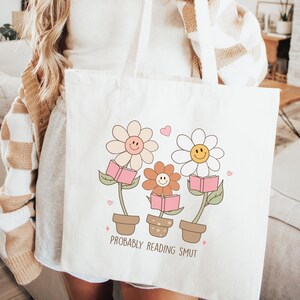 以下が含まれることがあります： 白いトートバッグに、本を持っている笑顔のデイジーの花が3つデザインされています。花の下には「PROBABLY READING SMUT」という文字が印刷されています。