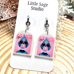 May include: A pair of dangle earrings featuring a cartoon Grinch character blowing a bubblegum bubble, wearing heart-shaped sunglasses, and set against a pink background. The word "Grinch" is printed above the image. The earrings are attached to silver hoops.