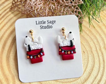 May include: A pair of dangle earrings featuring a white dog character resting in a red stocking with a white cuff, decorated with a string of colorful Christmas lights. The earrings are attached to a white display card labeled Little Sage Studio.