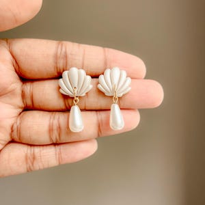 Peut inclure: Une paire de boucles d'oreilles blanches en forme de coquillage avec un pendentif en forme de goutte nacrée. Les boucles d'oreilles sont tenues par une main, mettant en valeur le design délicat et les accents dorés. Bijoux.