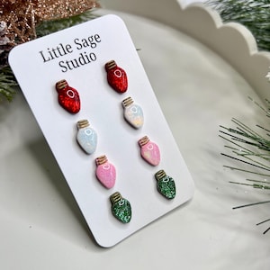 May include: A set of six colourful Christmas light bulb stud earrings. The earrings are red, white, light blue, pink, and green with gold-coloured bases. The earrings are displayed on a white card with the text "Little Sage Studio".