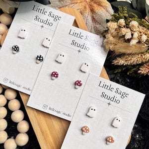May include: Three pairs of stud earrings on white cards, each with a ghost and mushroom design. The mushrooms have red, black, and brown caps. The cards are labelled "Little Sage Studio" with an Instagram handle. The earrings are displayed on a wooden tray.