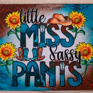 May include: A rectangular mat with a turquoise and brown background. The mat features the words "little MISS Sassy PANTS" in a mix of fonts and colors. Cowboy boots, a cowboy hat, sunflowers, and a bull skull are also included in the design.