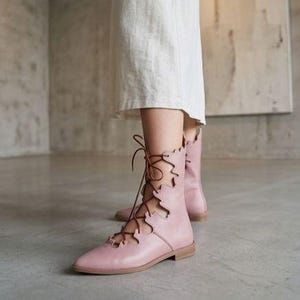 May include: Pale pink lace-up ankle boots with a pointed toe and a flat sole. The boots have a decorative scalloped edge and brown laces. The boots are worn with a white dress.