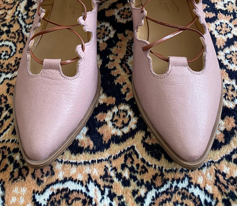 Puede incluir: Un par de bailarinas de color rosa p&aacute;lido con puntera y cordones marrones. Los zapatos tienen un dise&ntilde;o de borde festoneado y una suela marr&oacute;n claro. La imagen muestra un primer plano de los zapatos sobre una alfombra estampada.