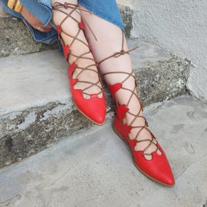 May include: Red leather flats with pointed toes and intricate brown lace-up detailing. The shoes feature cut-out designs along the sides and are shown on a concrete surface. The laces wrap around the ankles and legs.