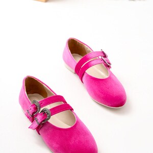 May include: A pair of pink velvet ballet flats with silver buckle accents. The flats have two straps across the top of the foot.