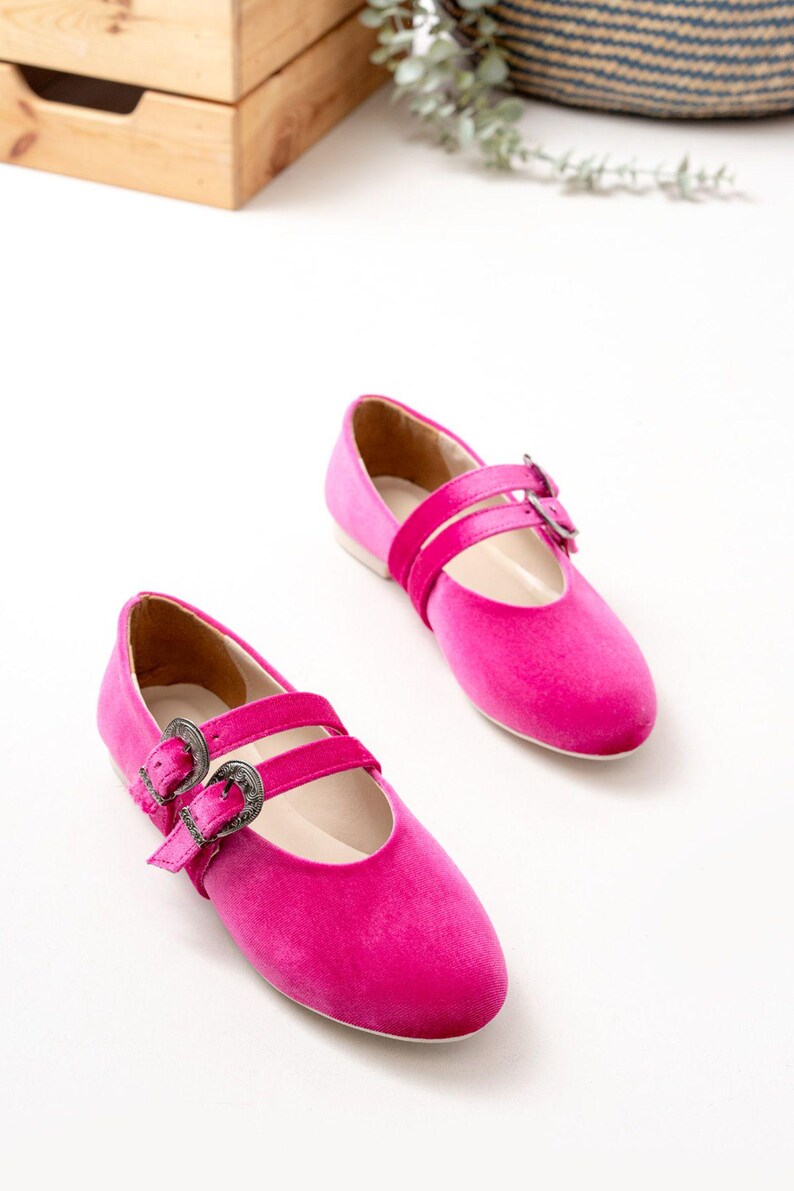 May include: A pair of pink velvet ballet flats with silver buckle accents. The flats have two straps across the top of the foot.