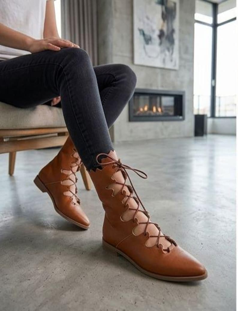 May include: Tan leather mid-calf boots featuring a pointed toe, a flat sole, and a decorative scalloped edge along the front opening. The boots are secured with thin brown laces that crisscross through metal eyelets up the front shaft.
