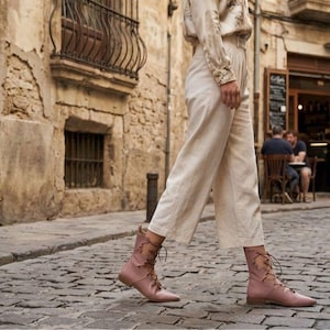 Puede incluir: Una persona caminando por una calle adoquinada con botas rosas con cordones. Las botas tienen punta y tac&oacute;n bajo. La persona tambi&eacute;n lleva pantalones color crema y una camisa de manga larga. El fondo muestra edificios antiguos y una cafeter&iacute;a.