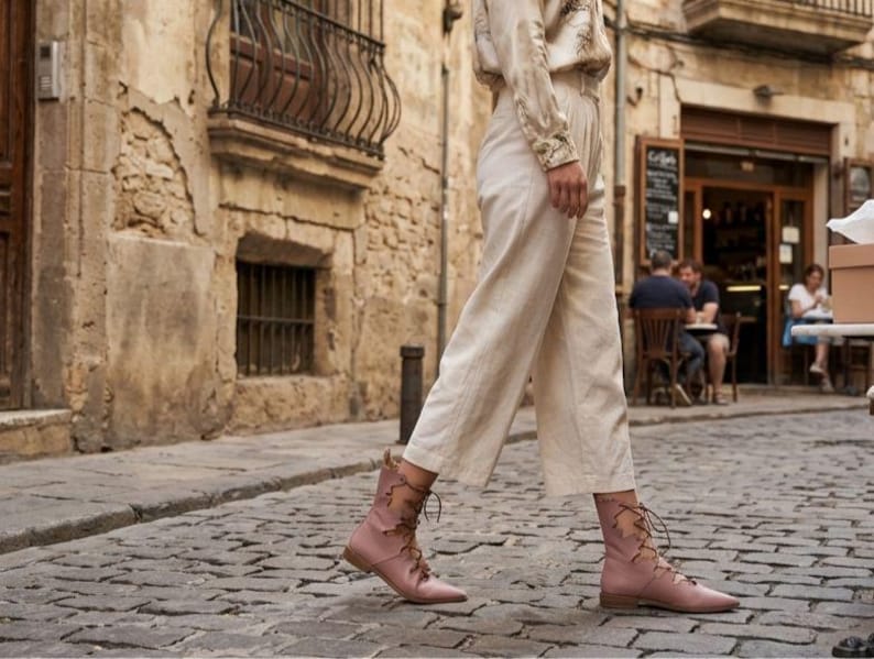 Puede incluir: Una persona caminando por una calle adoquinada con botas rosas con cordones. Las botas tienen punta y tac&oacute;n bajo. La persona tambi&eacute;n lleva pantalones color crema y una camisa de manga larga. El fondo muestra edificios antiguos y una cafeter&iacute;a.