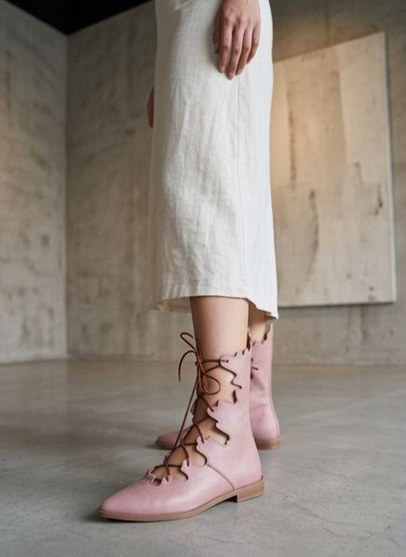 Puede incluir: Botines rosas p&aacute;lidos con cordones, punta afilada y suela plana. Las botas tienen un borde festoneado decorativo y cordones marrones. Las botas se llevan con un vestido blanco.