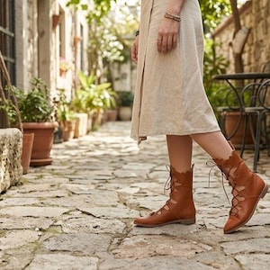 May include: A pair of tan leather lace-up boots featuring a scalloped edge design along the ankle and a flat sole. The boots are worn with a light-colored midi skirt, showcasing the intricate lacing detail that extends from the toe to the ankle.