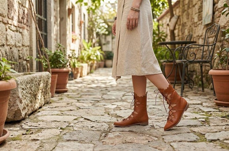 May include: A pair of tan leather lace-up boots featuring a scalloped edge design along the ankle and a flat sole. The boots are worn with a light-colored midi skirt, showcasing the intricate lacing detail that extends from the toe to the ankle.