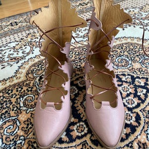 Puede incluir: Un par de botas de cuero rosa claro con cordones y un dise&ntilde;o &uacute;nico en forma de hoja en la parte superior. Las botas tienen cordones marrones y una punta puntiaguda. Las botas se exhiben sobre una alfombra estampada.
