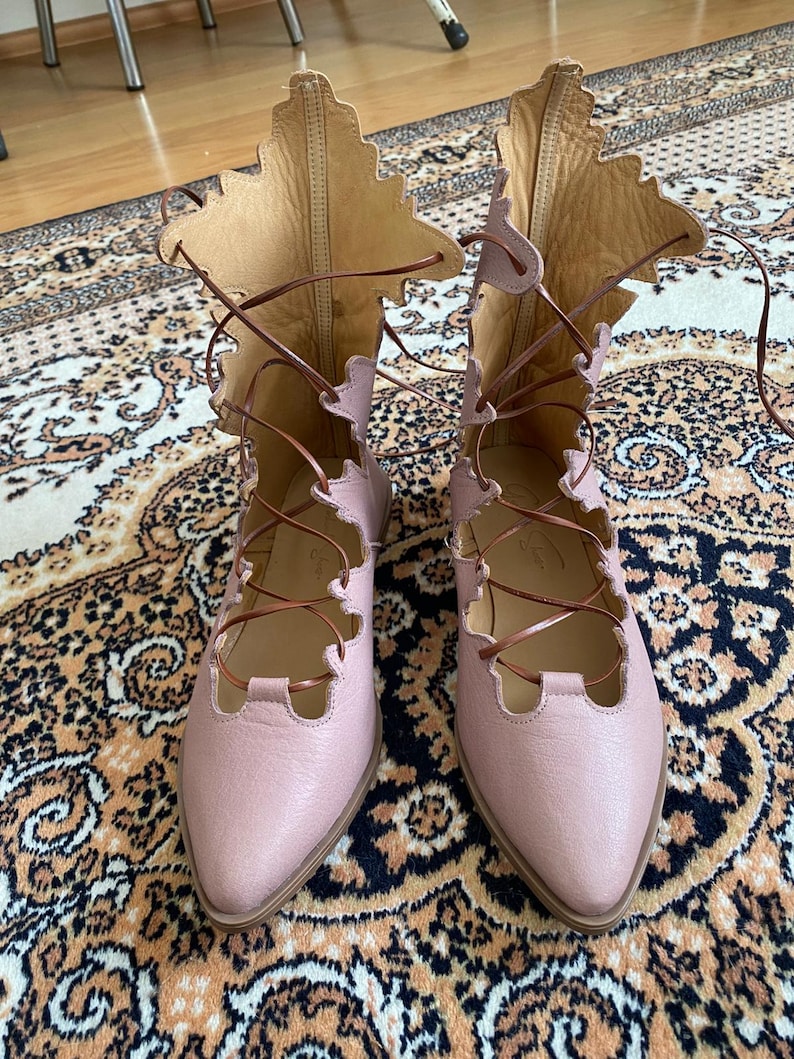 Puede incluir: Un par de botas de cuero rosa claro con cordones y un dise&ntilde;o &uacute;nico en forma de hoja en la parte superior. Las botas tienen cordones marrones y una punta puntiaguda. Las botas se exhiben sobre una alfombra estampada.