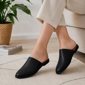 May include: A pair of black slip-on mules featuring a textured, perforated upper design with a closed, pointed toe and a flat sole. The footwear has a simple, backless construction that allows for easy entry and a clean, streamlined appearance.