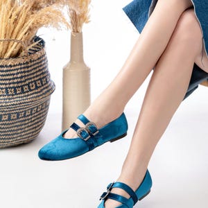May include: A pair of teal blue velvet flats with double buckle closures. The shoes have a pointed toe and a low heel.