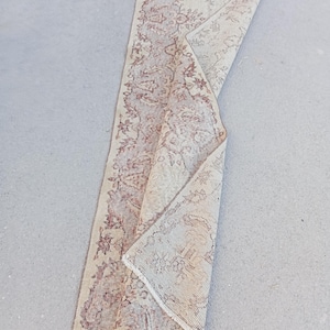 Pode incluir: Um tapete retangular creme com um padrão floral desbotado em tons de bordô e castanho. O tapete está parcialmente enrolado, revelando a parte inferior e a franja. O tapete é provavelmente feito de material tecido.