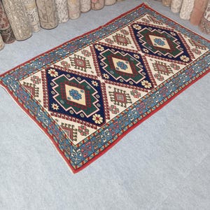 May include: Rectangular area rug with a cream-colored background and a repeating geometric pattern in navy blue, red, green, and beige. The rug has a red border and is surrounded by rolled-up rugs.