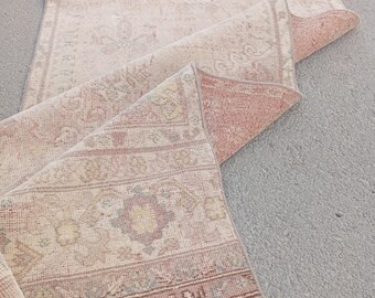 May include: A vintage-style rug with a faded, distressed appearance. The rug features a cream-colored central field with intricate floral and geometric patterns in muted shades of red, green, and brown. The edges are fringed, and the rug is partially rolled up.
