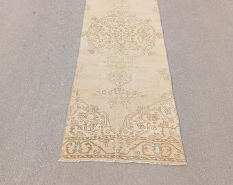 May include: A rectangular, beige-colored rug with an intricate, faded floral pattern. The rug has a distressed, vintage appearance, with a central medallion design and borders in shades of brown and teal. The rug is approximately 3 feet wide and 8 feet long.
