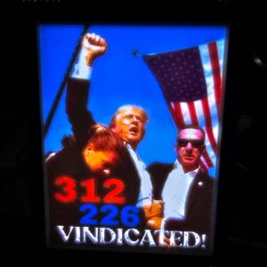 May include: A photo of a man in a black suit raising his fist in the air. He is standing in front of a crowd of people. The man is wearing a red and blue tie and a white shirt. The crowd is holding up signs that say "312 226 VINDICATED!" The man is standing in front of an American flag.