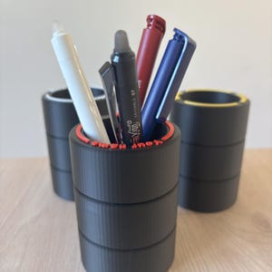 Puede incluir: Soportes para bolígrafos negros con borde rojo, que contienen bolígrafos de varios colores. Los soportes para bolígrafos son cilíndricos y apilados, con el texto "your name here" visible en el borde. El fondo es una superficie de madera clara.