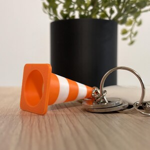 Op de afbeelding: Een miniatuur oranje en witte verkeerskegel sleutelhanger met een vierkante basis en een conische bovenkant. De verkeerskegel is bevestigd aan een zilveren metalen ring, waaraan verschillende platte ronde metalen sleutels zijn bevestigd.