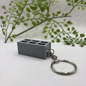 May include: A gray concrete block keychain with a silver keyring. The miniature block has six rectangular openings. The background includes green leafy sprigs on a white surface.