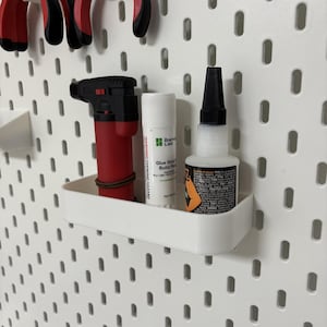 Puede incluir: Un panel perforado blanco con varias herramientas y suministros. Un soplete rojo y negro, una barra de pegamento y un frasco de adhesivo están en un estante blanco. Unas tenazas rojas y negras cuelgan encima.